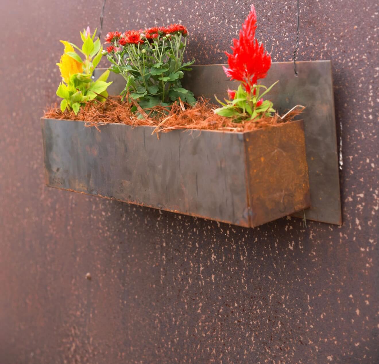 A handcrafted hanging Corten steel planter mounted on a wall, filled with bright red and yellow flowers.