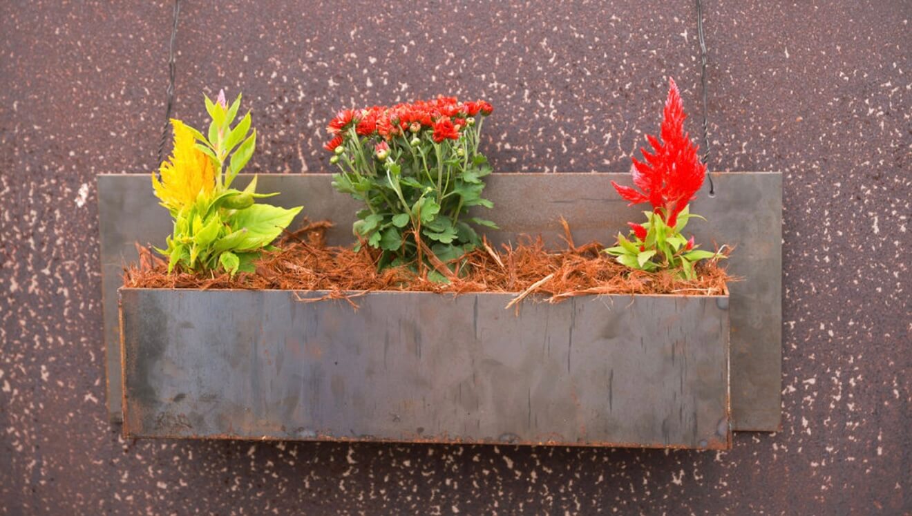 A front view of a rectangular hanging Corten steel planter showcasing red, yellow, and green plants against a rusted steel background.