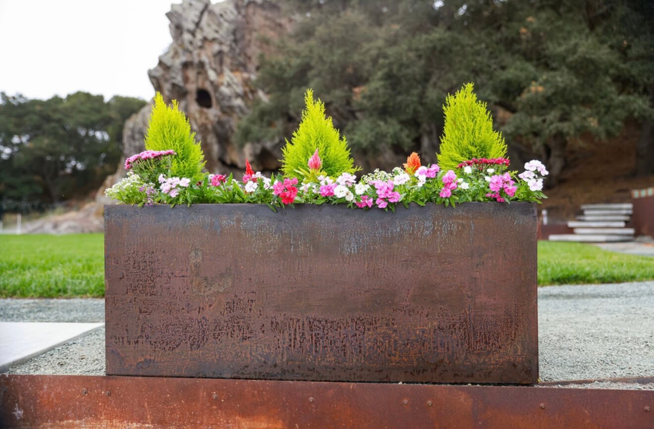 A long rectangular Corten steel planter filled with colorful flowers and green shrubs placed in a landscaped outdoor setting.
