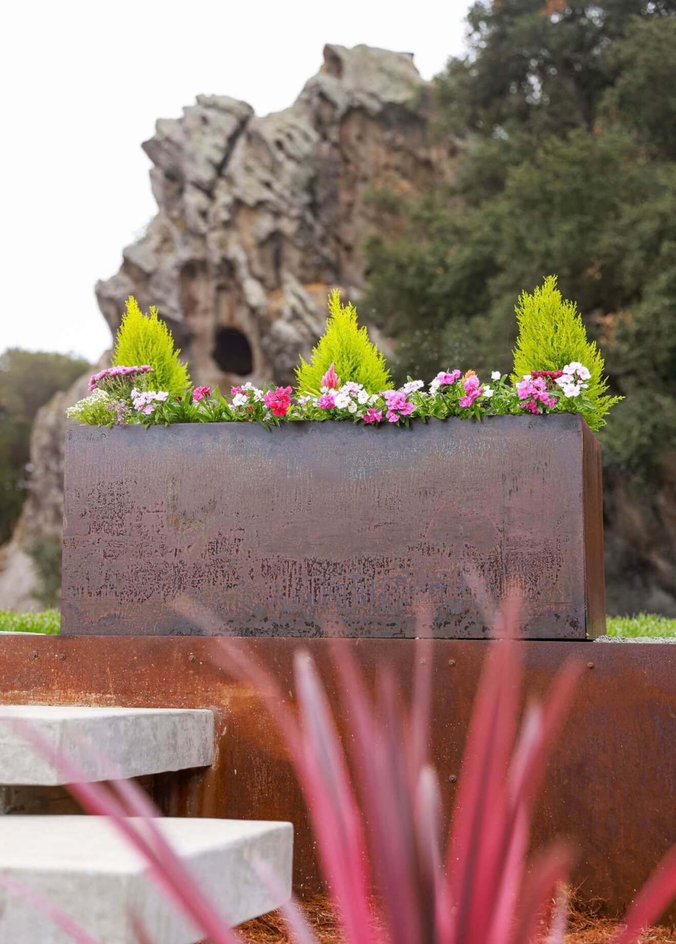 A large rectangular weathering steel planter surrounded by trees and greenery, featuring a rust patina and colorful garden plants.