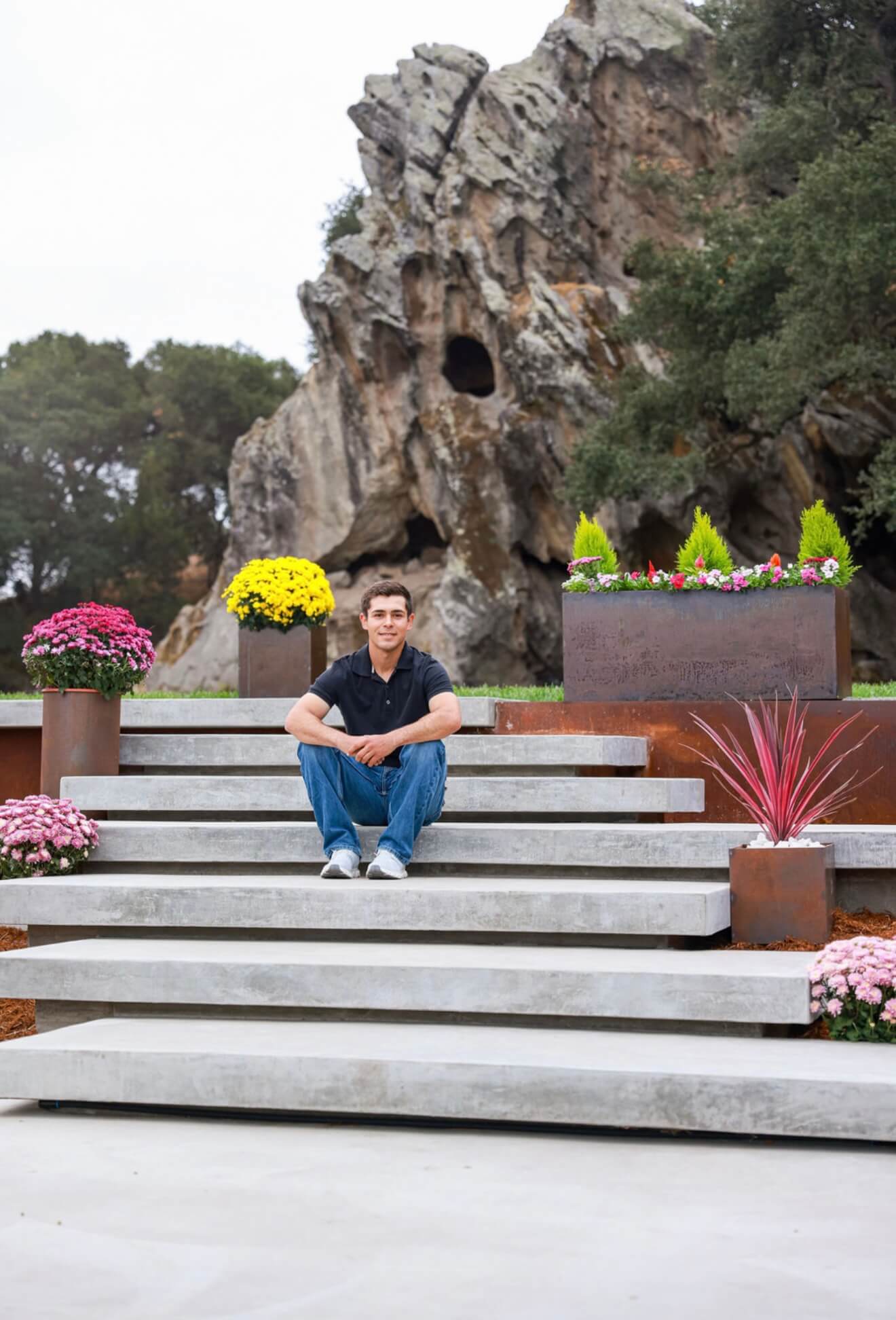 Moises Hernandez, fabricator and founder of Steel Root Design, sitting on concrete steps surrounded by Corten steel planters filled with colorful plants.